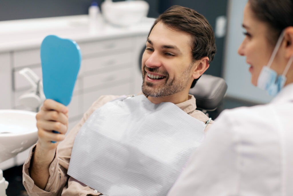 Happy male patient smiling while looking in mirror during dental appointment at clinic, being satisfied with result of procedure