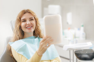 Happy young caucasian female at dental clinic holding mirror and smiling after professional whitening and teeth cleaning.