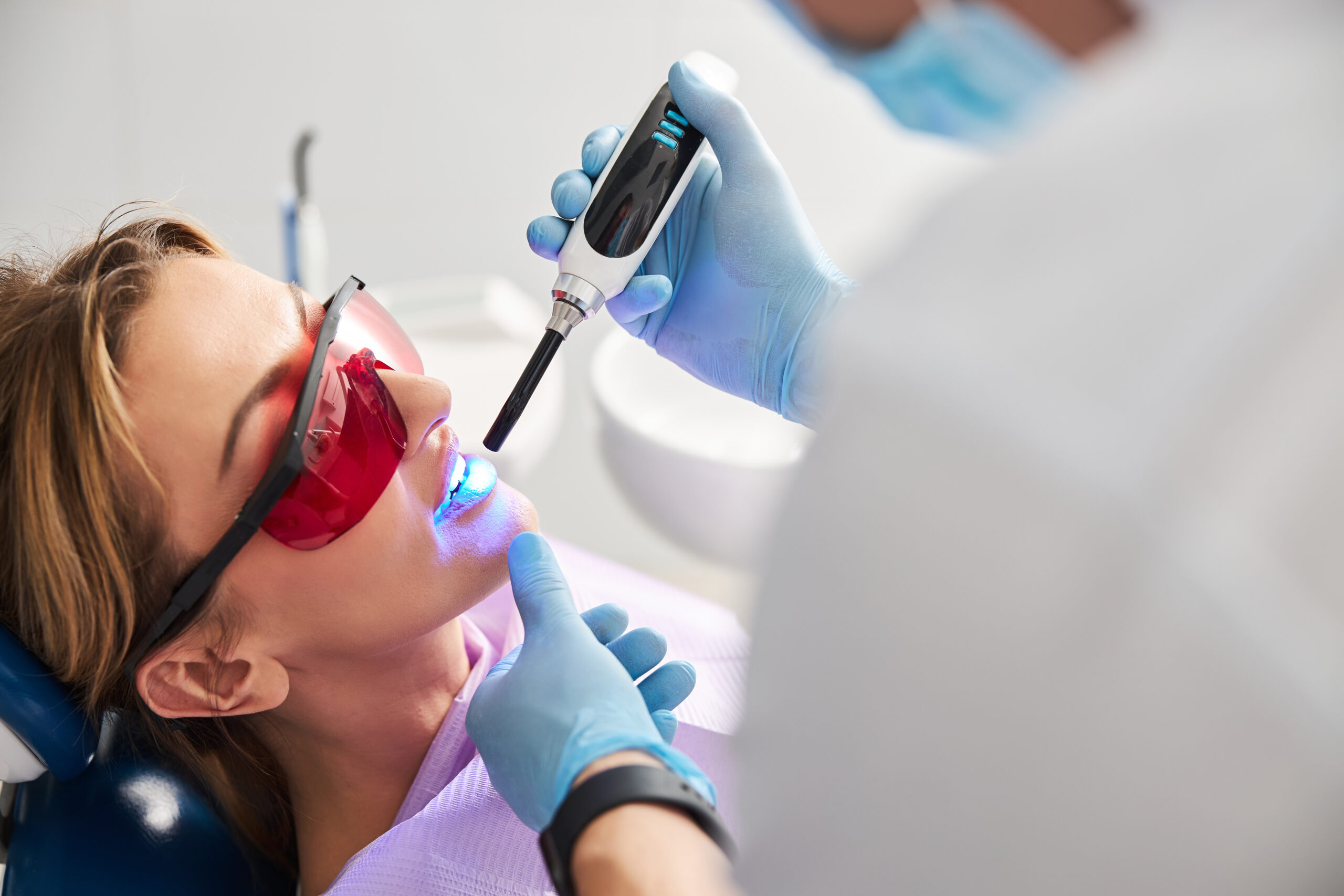 Medic shining with LED curing light on lady teeth for hardening of dental fillings while woman wearing red safety glasses