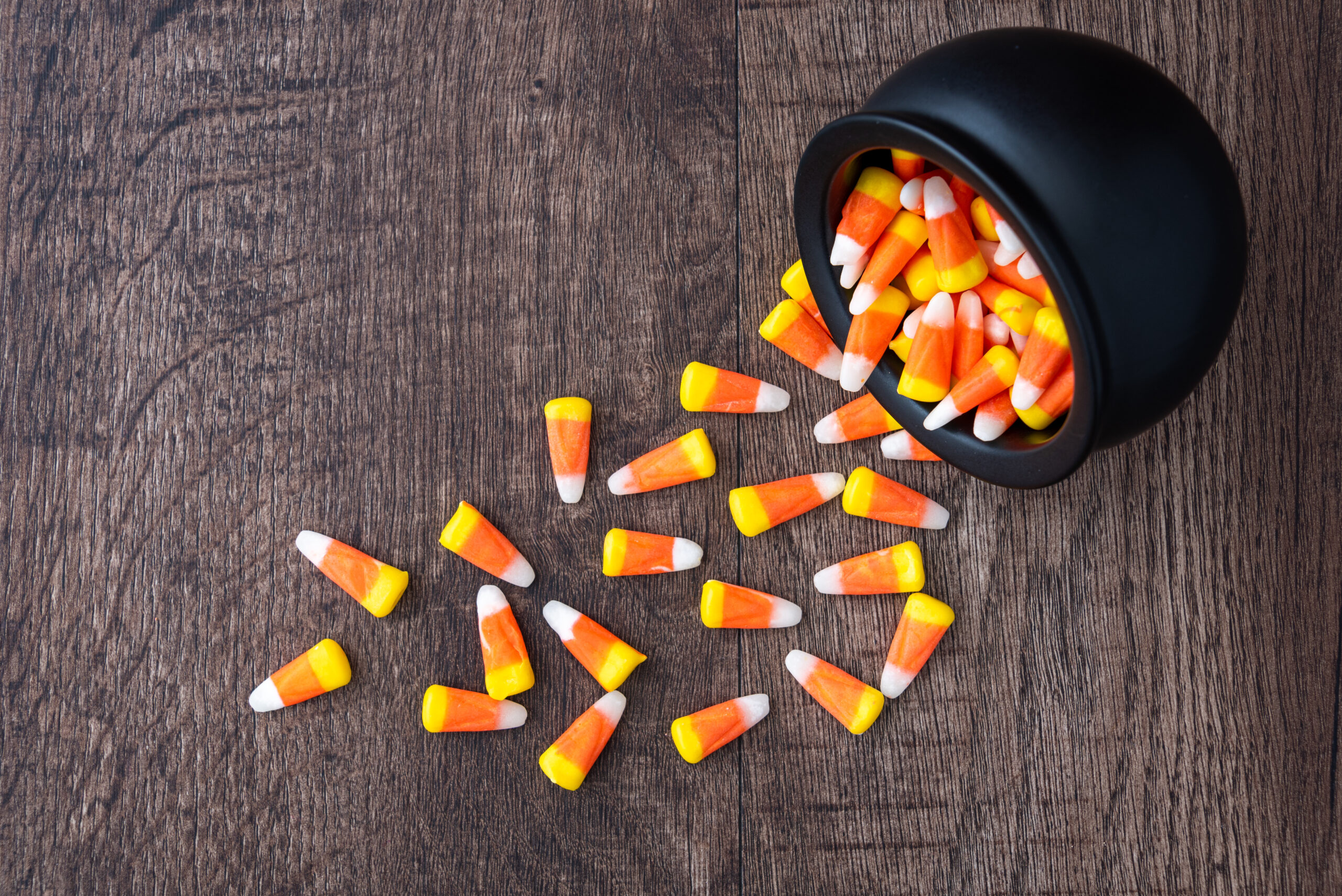 Black ceramic cauldron filled with holiday candy corn tipped over on a rustic wood background, spilling over onto the background