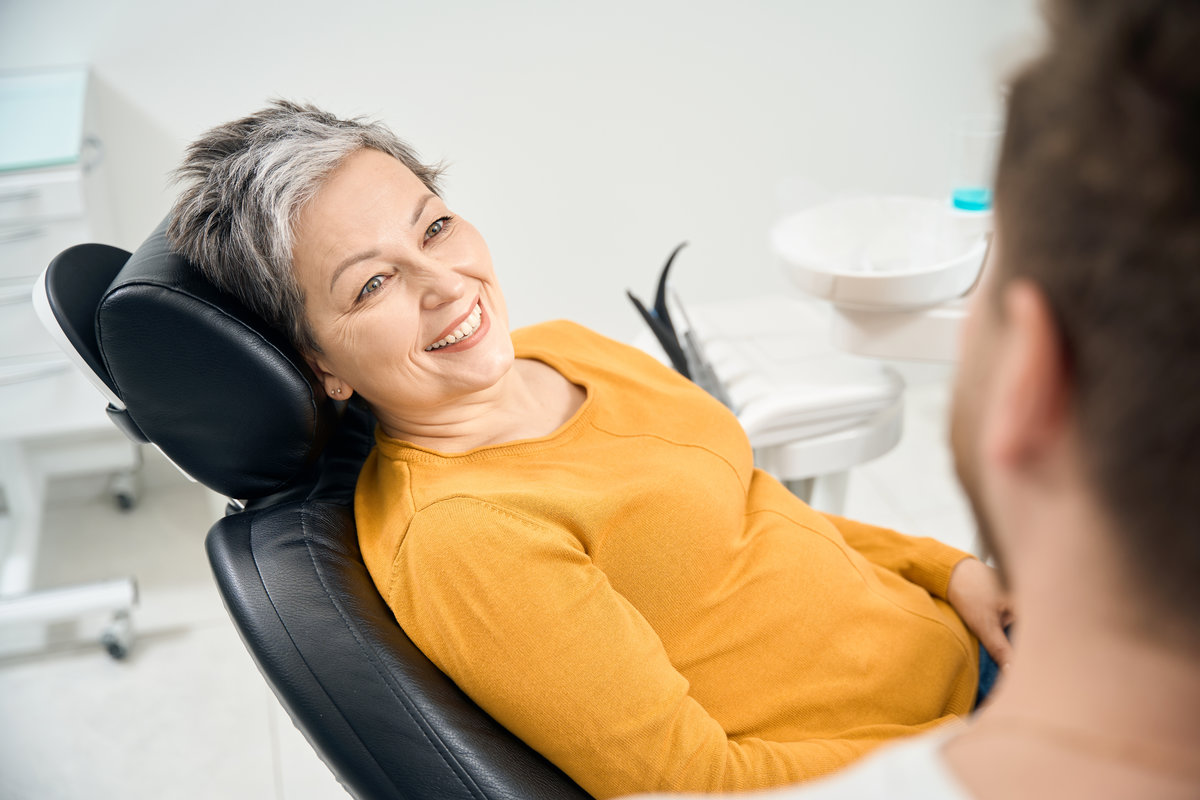 Stylish woman client in her 50 looking at her doctor stomatologist and smiling, satisfied with her healthy teeth and oral cavity, carefully listening to dentist recommendations