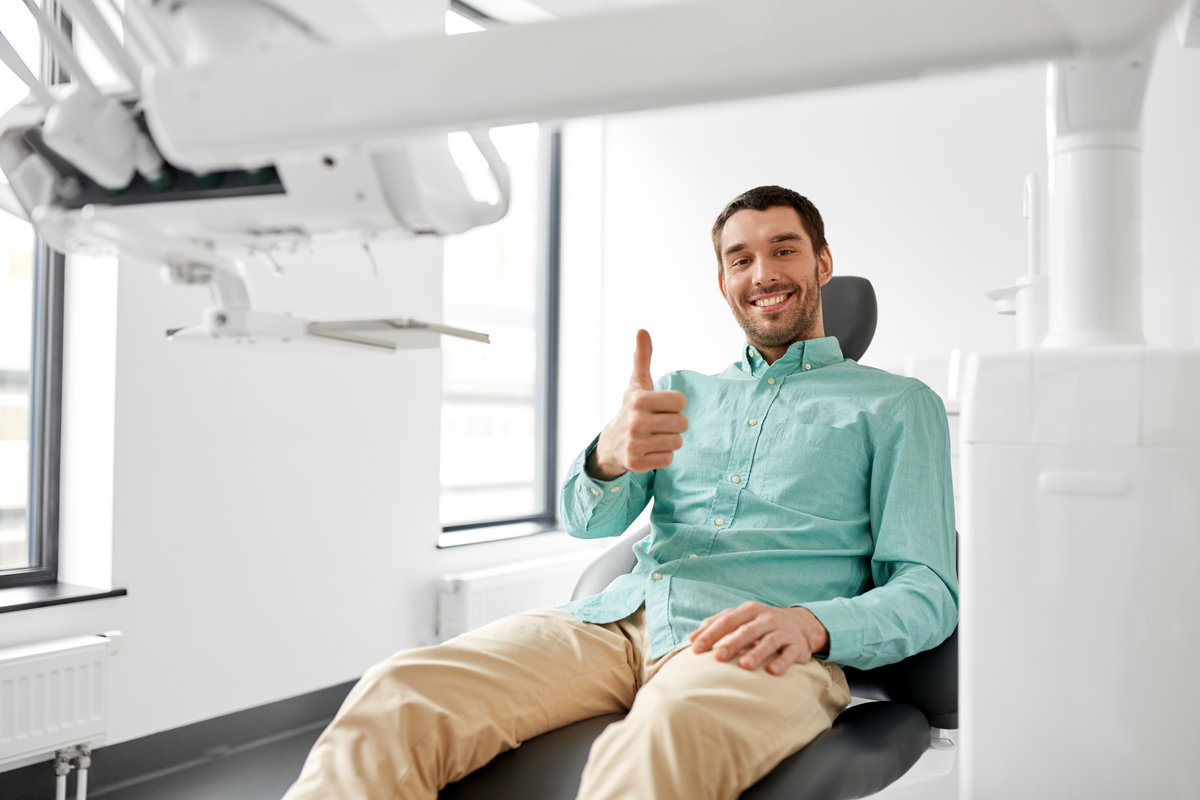 medicine, dentistry and healthcare concept - happy smiling male patient in chair showing thumbs up at dental clinic